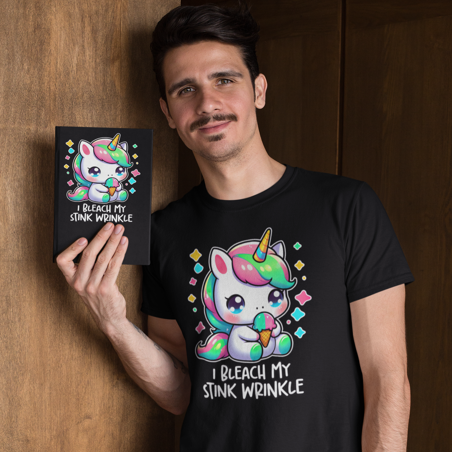 A person holding a black t-shirt with a colorful unicorn graphic and the text 'I Bleach My Stink Wrinkle' printed on it.