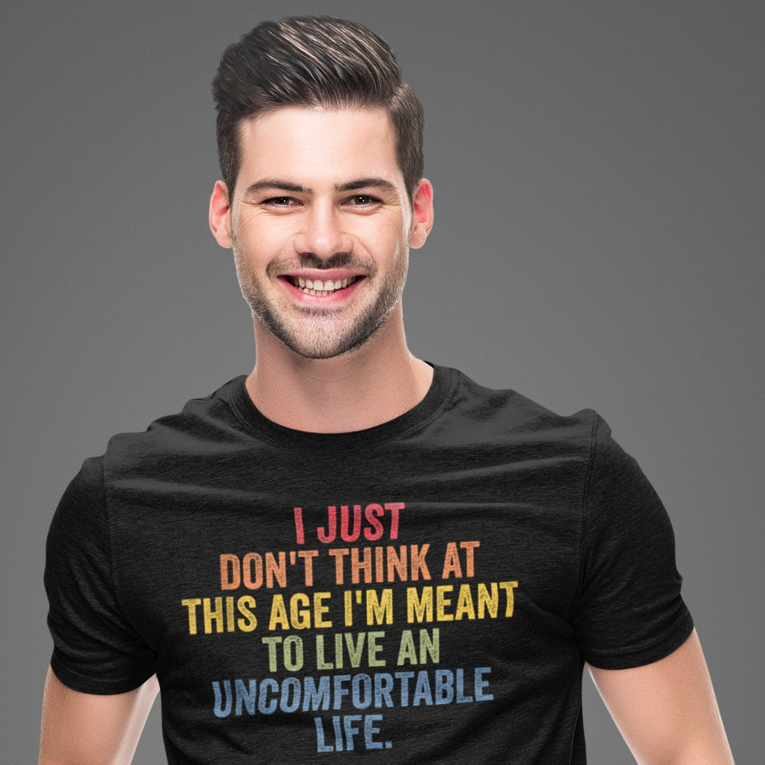 A man smiling and wearing a black crew neck t-shirt with a colorful text graphic that reads: 'I JUST DON'T THINK AT THIS AGE I'M MEANT TO LIVE AN UNCOMFORTABLE LIFE.'.